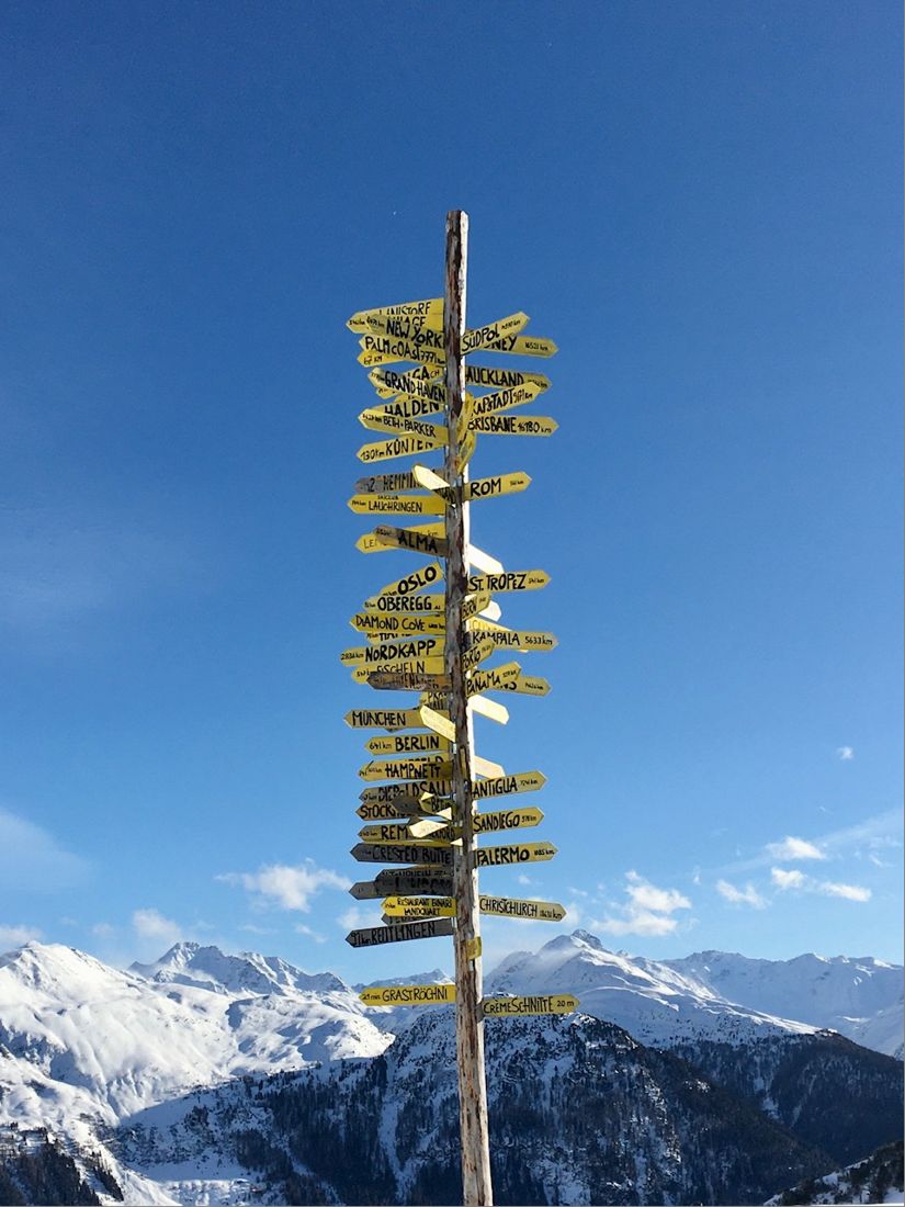 A wooden signpost covered in yellow arrows to places around the world, snowy Alpine peaks behind